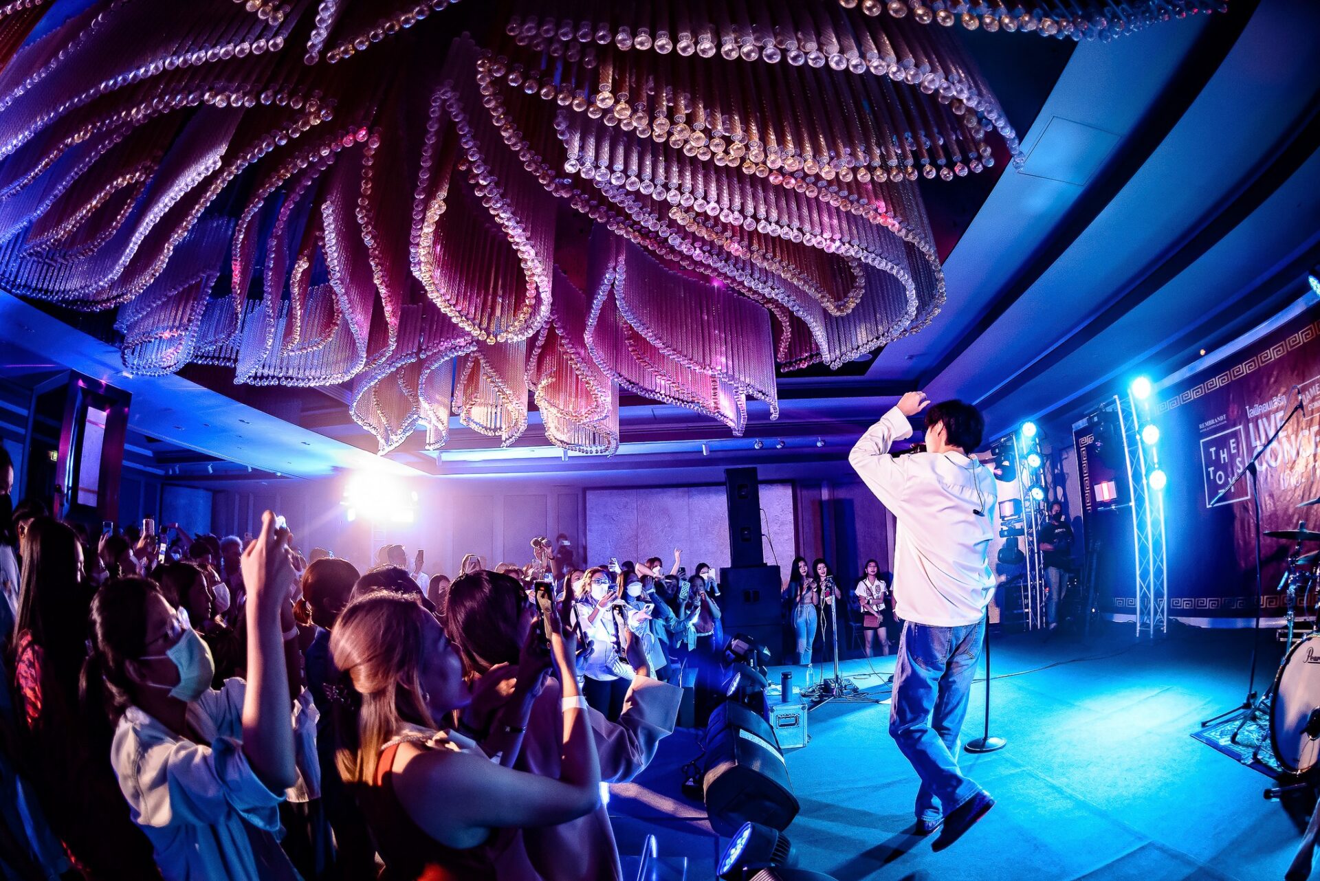 Live concert at Rembrandt Hotel Bangkok ballroom with singer performing on stage, vibrant lighting, luxury chandelier, and excited audience
