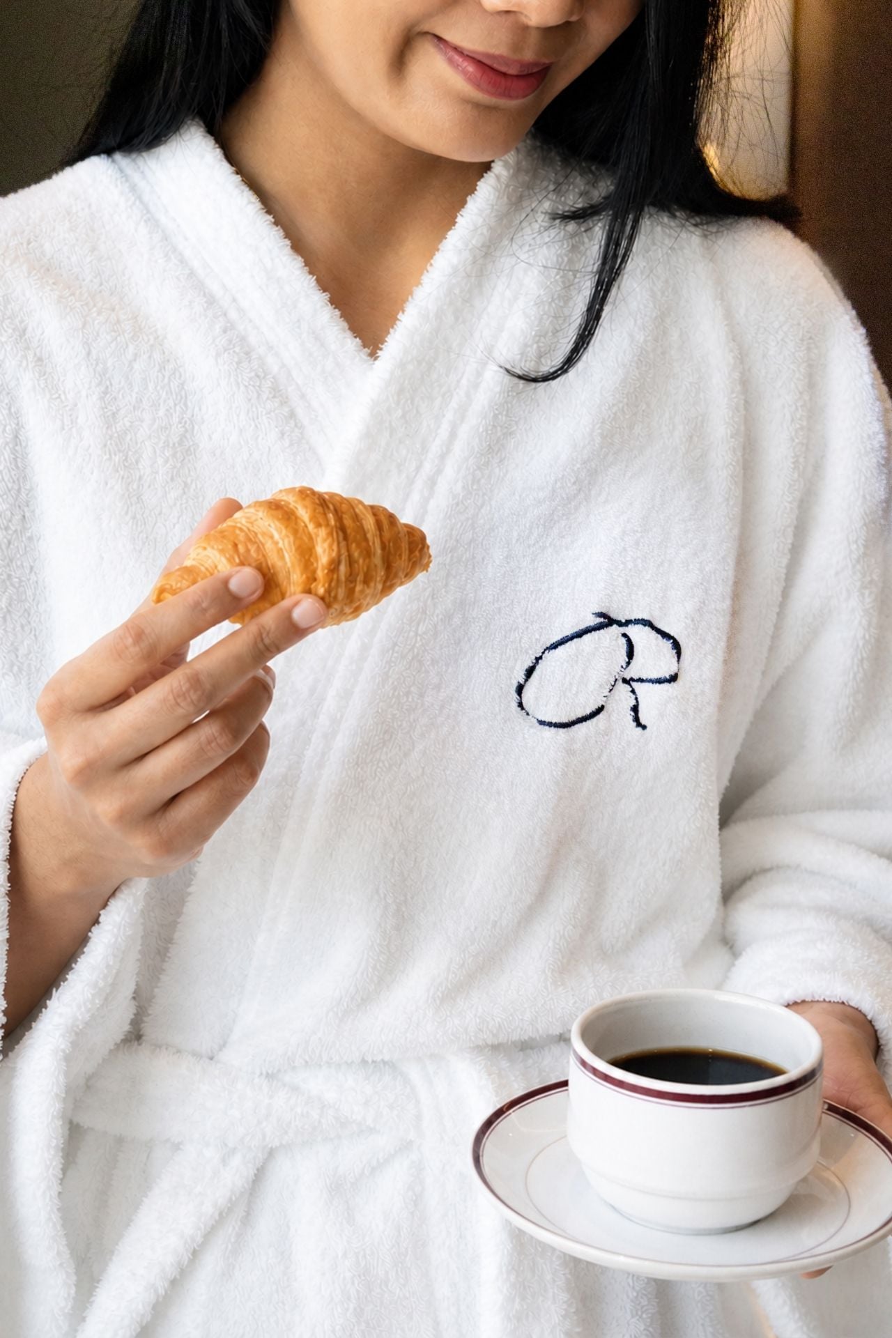 Woman enjoying croissant and coffee in hotel bathrobe